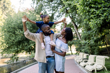 Young happy laughing African couple parents, spending time together and enjoying warm summer evening in park, walking with their little adorable daughter, sitting on shoulders of father
