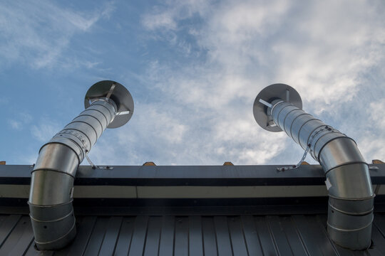 Two shiny metal supply and exhaust ventilation pipes mounted in the wall lined with siding under roof of metal profile. Pipes bend at right angles and rise up, ending in rain caps against cloudy sky
