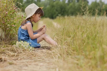 Foto op Canvas Schattig klein meisje in een hoed en een blauwe jurk op de landelijke weg met bloemen © Albert Ziganshin