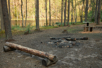 Beautiful landscape of bonfire on clearing by lake in forest. Campground with fire place, wooden benches and log on riverside. Perfect spot for camping. Hiking, tourism and active lifestyle