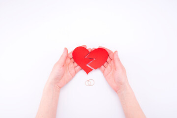 Fototapeta premium Female hands hold a broken heart on a white background with wedding rings. Relationship problems in marriage, divorce