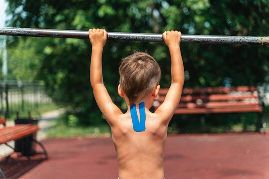 The young athlete pulls himself on the horizontal bar. Doing sports in the park. Teip on the back and neck, curvature of the spine, scoliosis, back problems