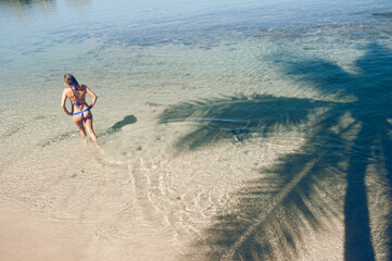 Femme en bikini de dos qui marche sur la plage, avec une ombre de cocotier à la surface de l'eau....