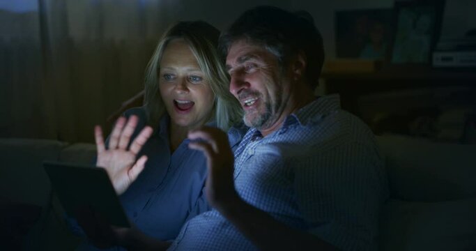 A happy mature couple is making a selfie or video technology call to their relatives with a tablet while sitting on a sofa in living room at home at night. Concept: technology, family, connection