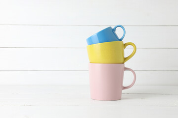 Stacked blue, yellow cups and pink coffee mug on white wood background with copy sapce.