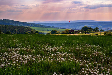 Idyllic spring countryside with blooming meadow, small homestead, hill, valley, floating red hot...