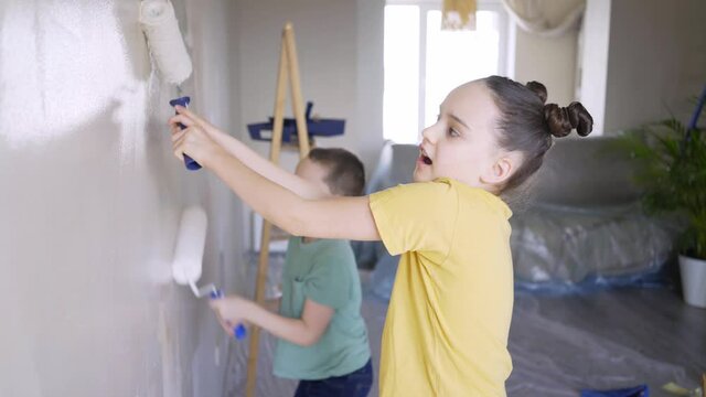 excited children colour new apartment wall into white holding rollers standing on floor with plastic cover against furniture during self isolation close view