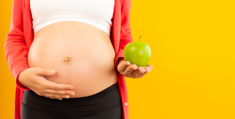 Happy pregnant young asian mother touch belly and holding fruit or green apple. Apple has vitamin, good for fetus, baby and pregnant woman with isolated on yellow background, copy space. Health care