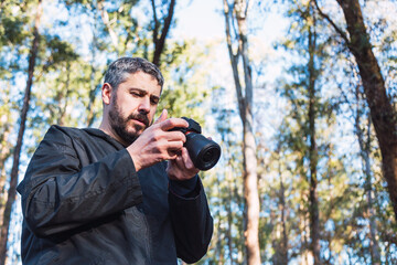A man checks his camera. He has a beard, and gray hair, about 40 years old, and walks in a park with many trees in the background.