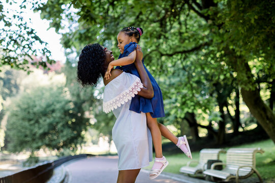 Young beautiful African woman mother in light blue dress playing with her little cute daughter in the park, having fun and lifting the baby up. Happy motherhood concept