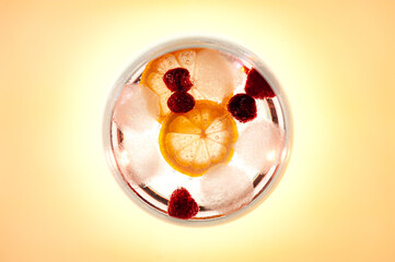 Top shot of a Gin and Tonic drink with raspberry’s and a slice of lemon. On yellow background with backlit.