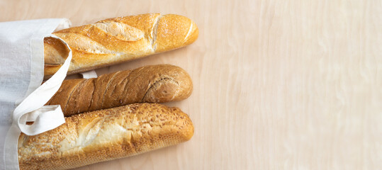 Fresh, fragrant baguettes made of rye and wheat flour, with sesame seeds, in a canvas bag, on a light wooden background. Banner. Ecological concept of the crop. Top view, close-up, selective focus.