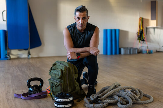 Motivated boot camp instructor kneeling with gym equipment in gym hall. Dumbbells, rope, sandbag on wooden floor. Portrait of lebanese instructor for gym concept.