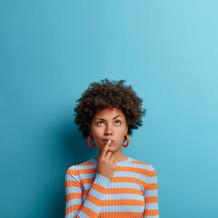 Vertical shot of thoughtful dark skinned woman has idea in mind, keeps finger on lips, looks pensively above, tries to decide how to act, poses against blue background, blank copy space above - obrazy, fototapety, plakaty