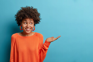 Let me introduce this. Cheerful friendly curly haired pretty woman with toothy smile, raises palm over blank copy space on blue background, advertises product on sale, dressed in orange jumper