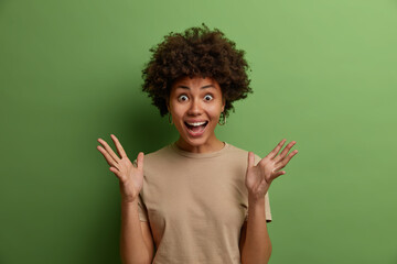 Joyful very excited cheerful African American woman happy to receive awesome present, raises hands and gestures actively, cannot believe her huge success, dressed in casual wear isolated on green wall