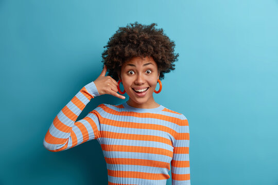 Give me call sometimes. Cheerful satisfied Afro American woman makes call me gesture, keeps connection and communication, poses on blue background, dressed in striped jumper. Sign and symbols