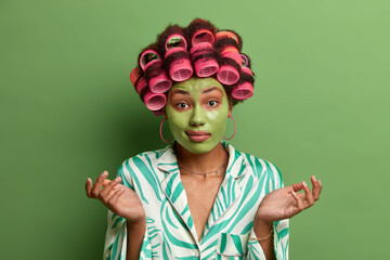 Easy tips for making hairstyle. Confused female model spreads hands sideways with doubt, uses hairdressing tools, spends free time in salon, applies green nourishing mask on face, dressed casually