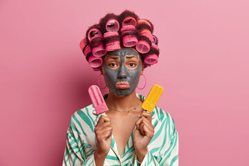 Displeased woman applies clay mask for skin care, wears hair curlers for making hairstyle, sad as cannot eat frozen ice cream containing much calories, dressed in casual gown, feels very upset