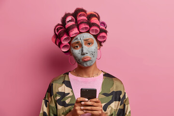 Sad displeased woman purses lips, doesnt get message from boyfriend uses mobile phone for surfing in social networks feels bored during beauty procedures wears hair curlers expresses negative emotions