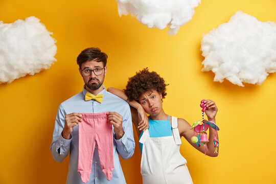 Bored sad expectant parents await for baby, buy clothes and crib mobile for newborn, pose togehter against yellow background, fluffy clouds above head. Future father and mother prepare for parenthood