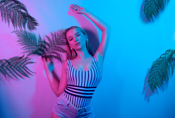 A model in neon poses. Fashion photo of a girl in a swimsuit and shorts in pink-blue light. Girl on a background of tropical leaves summer photo. Beautiful blonde