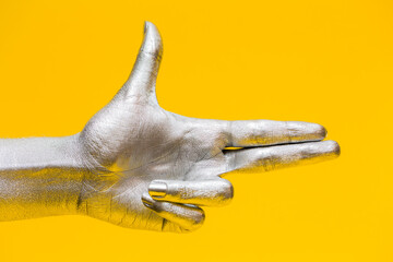 Fototapeta premium Elegant female hand with a silver paint on it shows gesture bang bang isolated on a yellow background. Free space for text