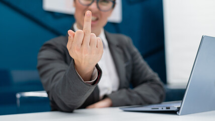 Angry business woman in a suit shows a middle finger while sitting at a desk. Annoyed female office worker showing a fuck you gesture. Professional burnout.