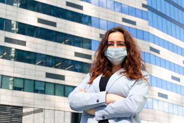 Caucasian business woman wearing protective mask stands with arms crossed. Blurred office building in the background. Copy space for your text. New Normal Theme. 