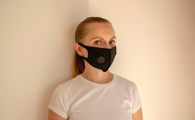 Young blond Caucasian woman in white t-shirt wears black reusable face mask. Protection against viruses and infections, light beige background