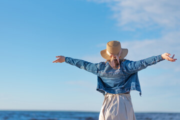 On the beach by the sea, a woman with her arms wide open, dressed in a beige wide dress, denim jacket and straw hat.