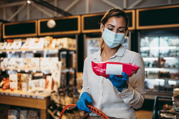 Beautiful young and elegant woman with face protective mask and gloves buying healthy food and drink in a modern supermarket or grocery store. Pandemic or epidemic lifestyle and consumerism concept.