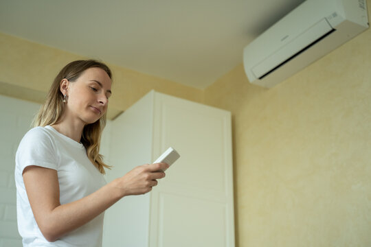 Woman in her modern apartment in living room. She turns on the air conditioner from the remote control. Climate control at home with split system. - Powered by Adobe