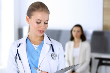 Doctor woman filling up medication history record while standing in emergency hospital office with patient in queue at background. Physician at work, portrait shoot. Medicine and health care concept