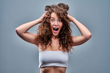 Portrait of amazed young woman with curly hair posing over grey background, keeping palms on her...
