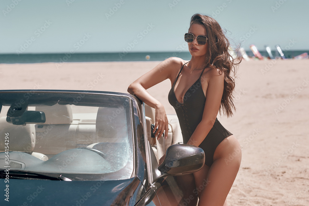 Wall mural Sexy tanned caucasian woman with wavy brunette hair standing at beach leaning on black cbriolet car on a sunny warm day. Fashion model in black swimsuit