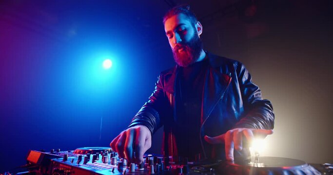 Authentic hipster dj rocking the party up. Bearded disc jockey working in a nightclub, composing a dance music list, spotted by red and blue lights - nightlife concept 4k footage