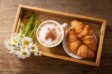 cup of cappuccino , croissants and daisy flowers on a wooden tray