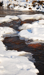 brown river water flows over stones with ice and snow in sunny winter day.