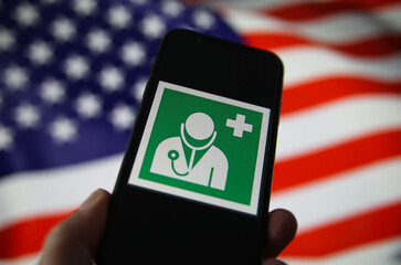 Viersen, Germany - July 9. 2020: View on isolated mobile phone screen with international health and medical doctor symbol. Blurred waving american flag background. (Focus on head of doctors symbol)