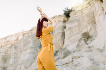 Young beautiful fashionable woman model posing on the background of sand quarry outdoors, looking away, wearing stylish yellow overalls and holding arms raised. Female fashion concept