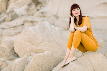 Summer lifestyle fashion image of pretty woman wearing stylish yellow overalls, sitting on the sand stone in quarry, enjoying moment and looking at camera.