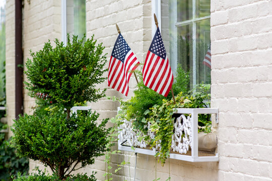 Old colonial style home with white painted bricks is adorned with American flags for the Fourth of July (Independence Day). Trees and green plants on the window planters.