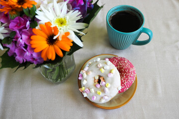 two doughnuts with pink icing and a white one with marshmallows on a brown round plate with coffee in a turquoise mug and a bouquet of flowers in a glass vase on a beige background side view
