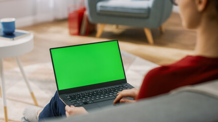 Young Woman at Home Works on a Laptop Computer with Green Mock-up Screen. She's Sitting On a Couch in His Cozy Living Room. Over the Shoulder Shot © Gorodenkoff