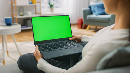 Young Woman at Home Sitting on a Couch Works on a Laptop Computer with Green Mock-up Screen. Girl Using Computer, Browsing through Internet, Watching Content, Chatting in Social Networks with Friends.