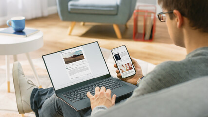 Young Man at Home is Using Laptop Computer, Srufing the Net, Watching News, while Holding...