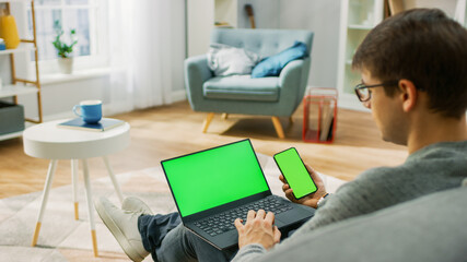 Young Man at Home Works on a Laptop Computer with Green Mock-up Screen, while Holding Smartphone...