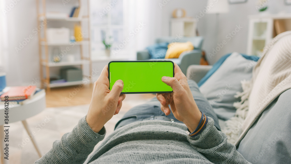 Poster man at home lying on a couch using smartphone, holds it horizontally in landscape mode. playing vide