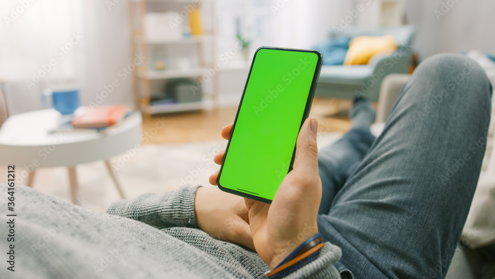 Poster man at home lying on a couch using smartphone with green mock-up screen. over the shoulder camera sh